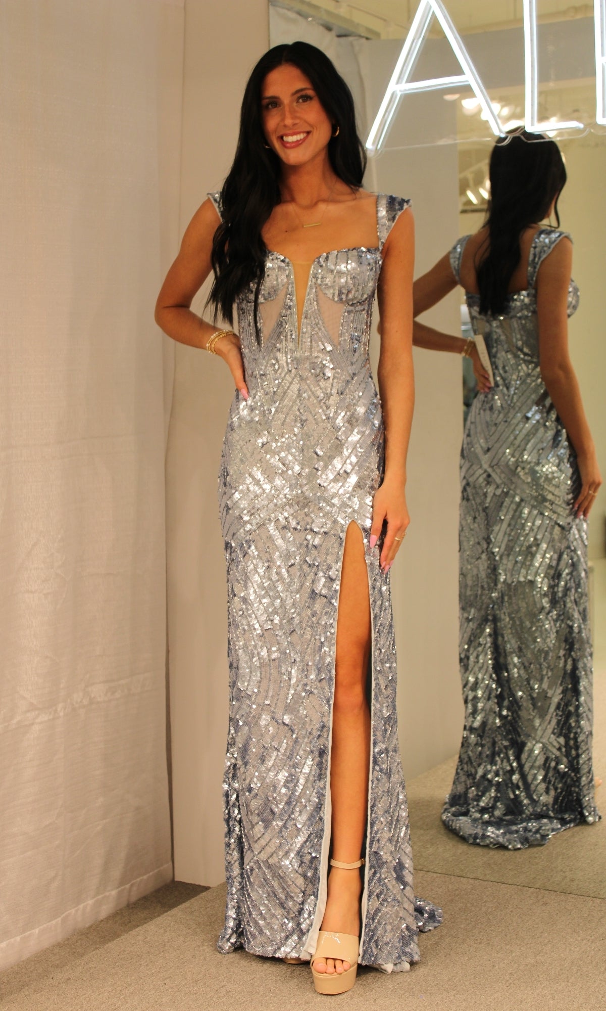 A woman in the Cap-Sleeve Long Sequin Prom Dress 1173 with a thigh-high slit and strappy heels stands smiling in front of a mirror.